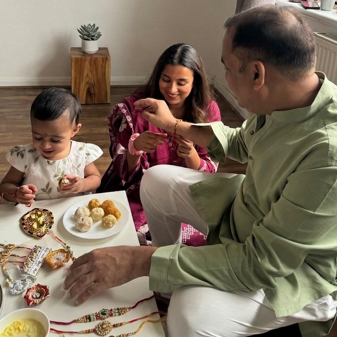 A man and a young child are celebrating Rakhi with a woman sitting in the background, all gathered around a table filled with sweets and decorated Rakhi bracelets in a home setting.